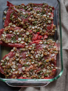 Raw skirt steak marinating in raw chopped onions and herbs in a glass dish.